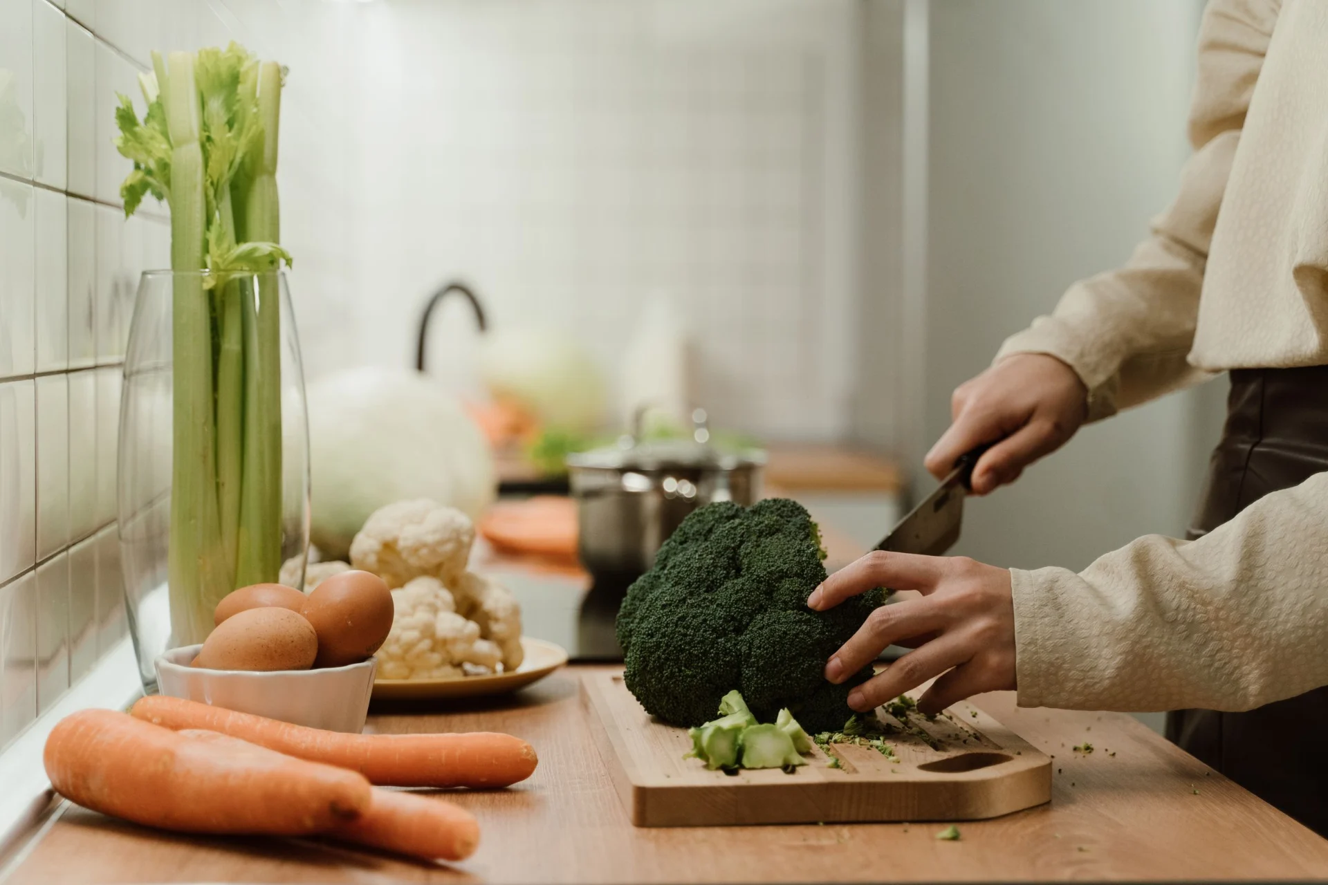 Person chopping broccoli on a wooden cutting board in a kitchen with various vegetables and eggs nearby.