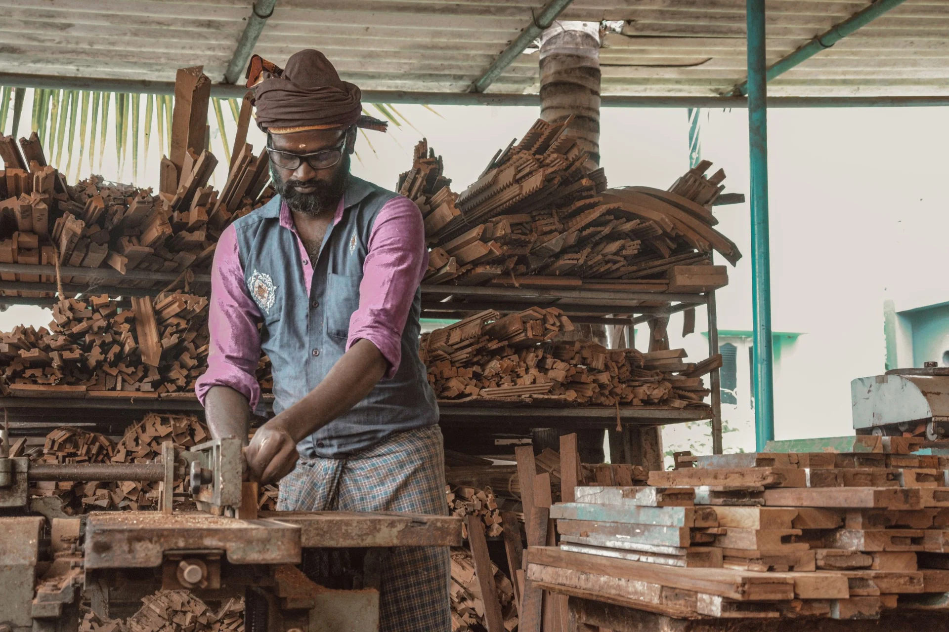 A man in traditional clothing works with wood in a workshop surrounded by piles of wooden planks.